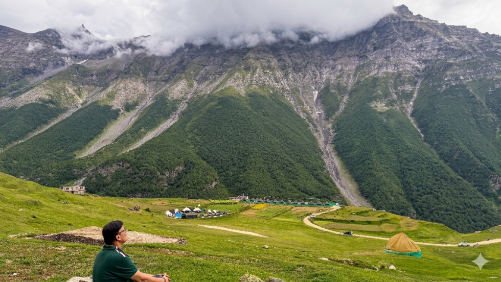 Person sitting on a grassy hill overlooking tents and green valley with mist-covered Himalayan mountains in Sural Bhatori, Pangi Valley