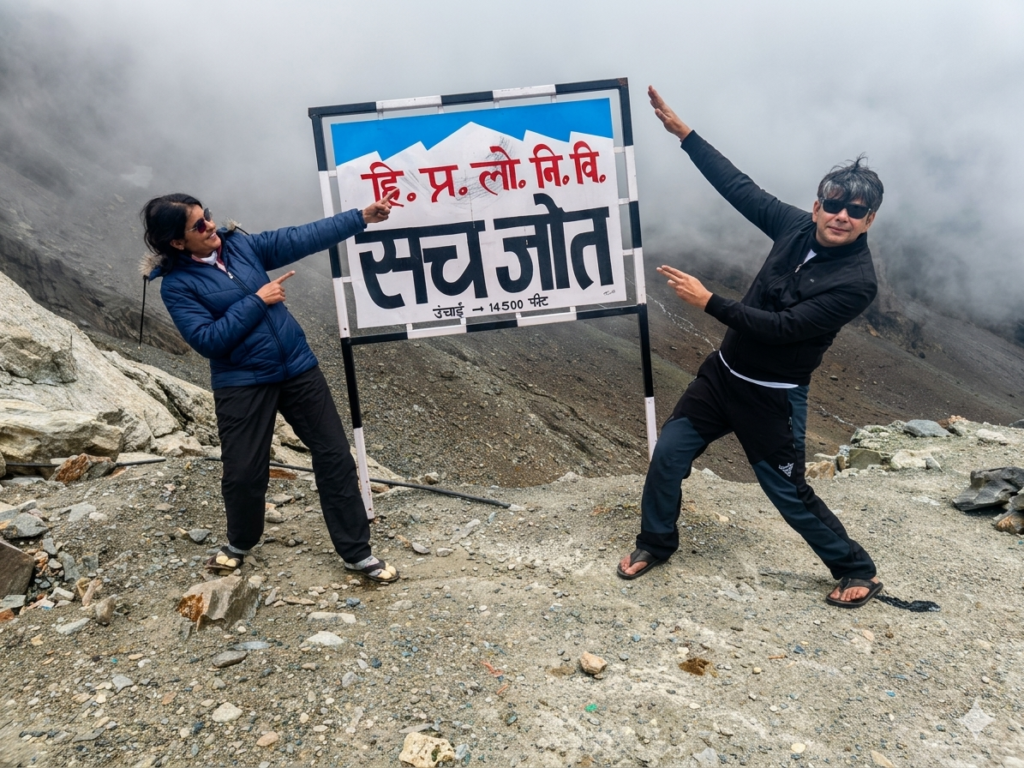 Two people posing near a signboard at Sach Pass with rocky terrain and foggy mountains in the background.