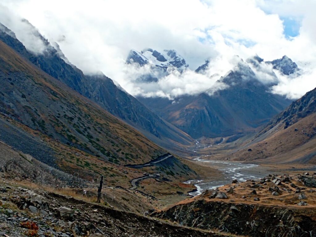 A deep mountain valley with a river running through the center, partially covered by low-hanging white clouds.