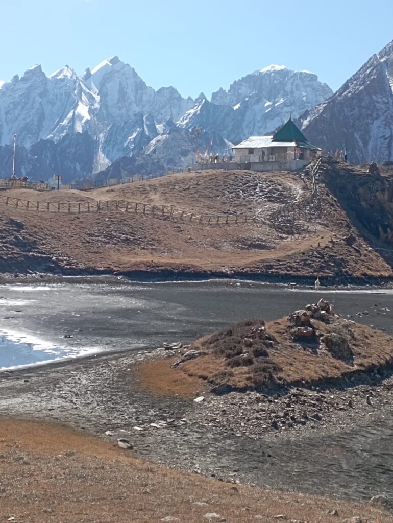 A small stone temple with a green roof situated on a rocky ridge against a backdrop of jagged snowy mountains.