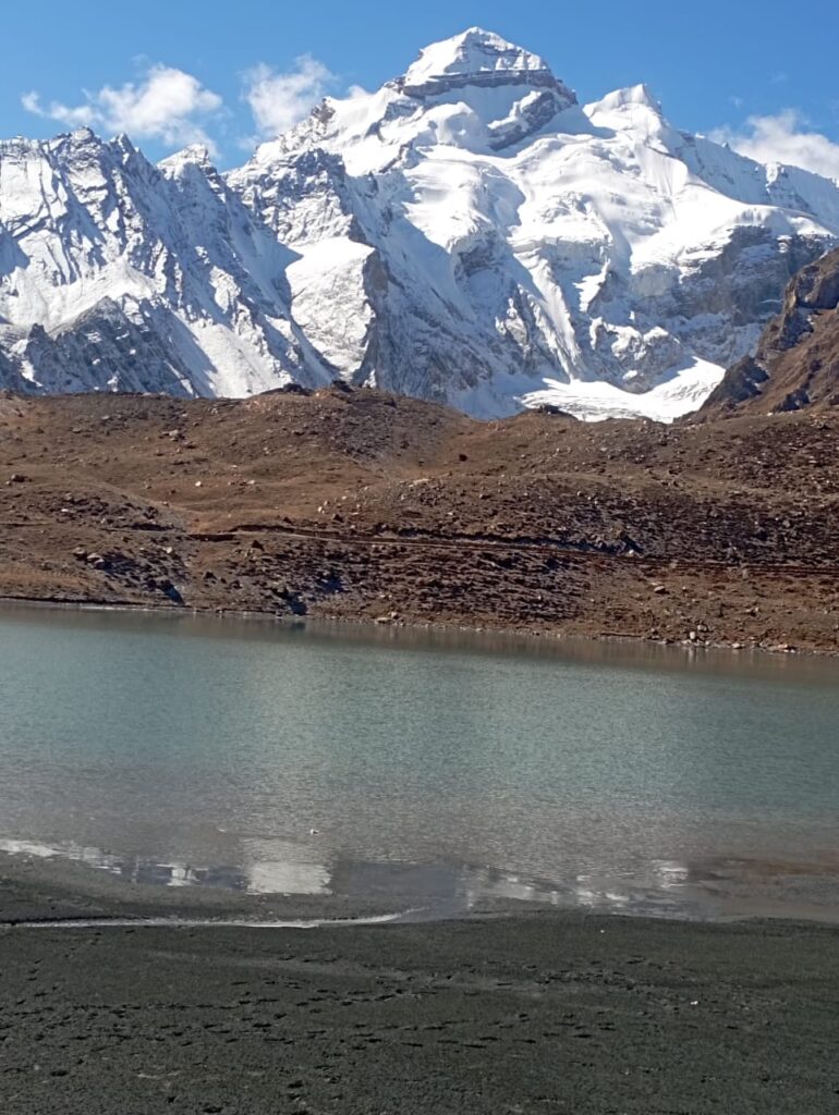 A calm high-altitude lake reflecting a snow-capped mountain peak in the background.