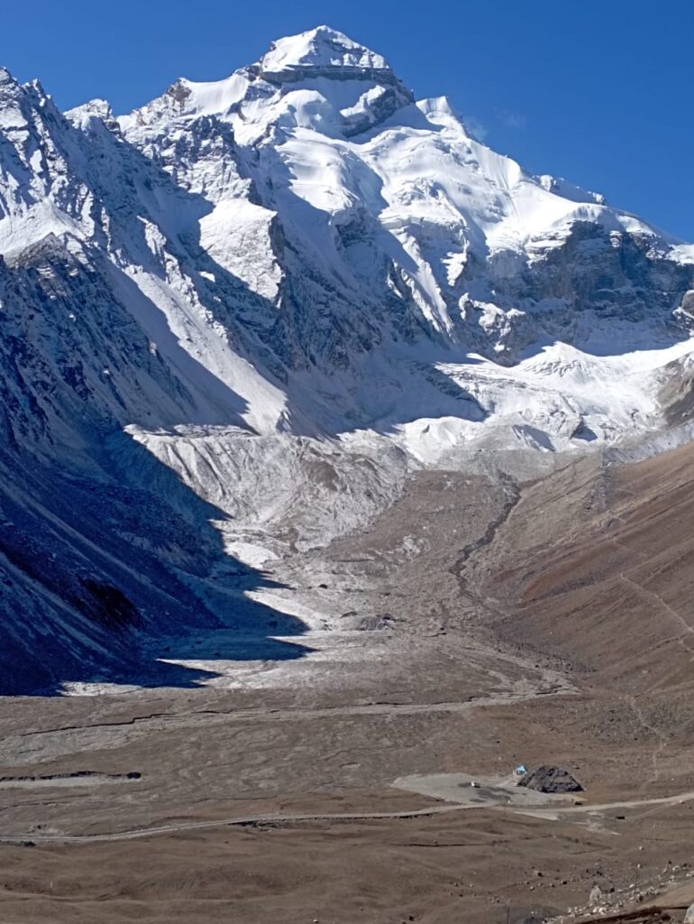 A towering, snow-covered mountain peak under a clear blue sky with a rocky valley floor in the foreground.