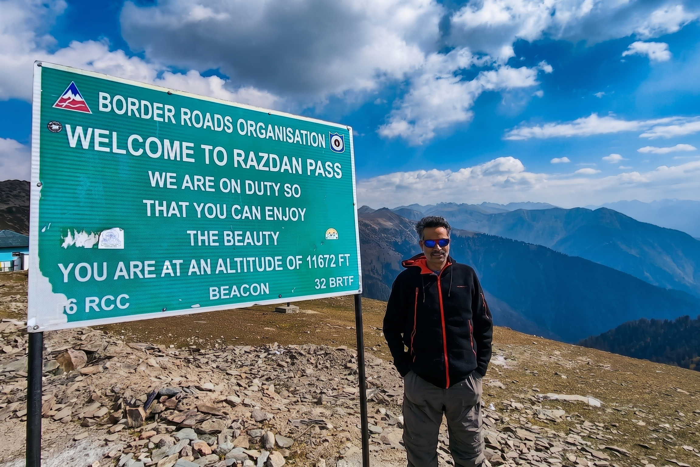 A traveler in sunglasses standing next to a large green Border Roads Organisation sign welcoming visitors to Razdan Pass at an altitude of 11,672 feet, with a backdrop of the sweeping Gurez Valley mountains under a blue sky.