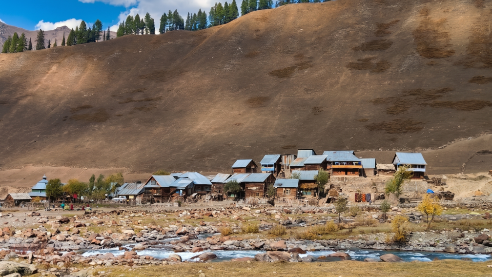 A scenic view of a small village with wooden houses and tin roofs nestled along a rocky, fast-flowing blue river at the base of a massive, barren mountain slope in Gurez Valley.