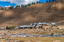 A scenic view of a small village with wooden houses and tin roofs nestled along a rocky, fast-flowing blue river at the base of a massive, barren mountain slope in Gurez Valley.