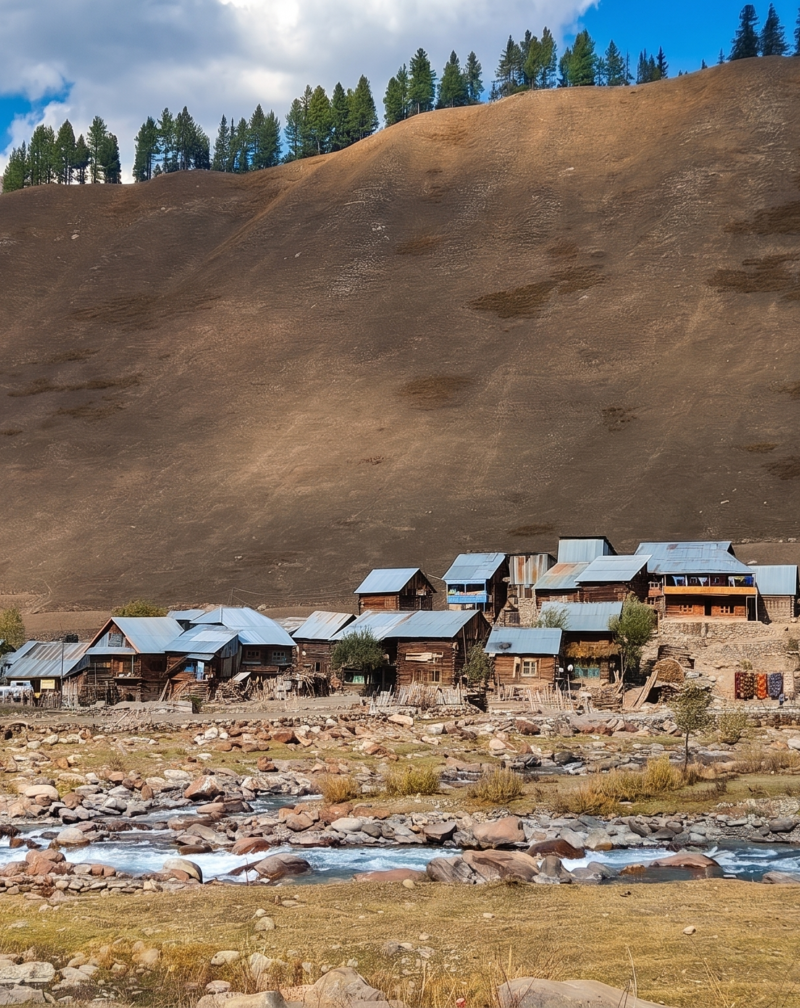 A scenic view of a small village with wooden houses and tin roofs nestled along a rocky, fast-flowing blue river at the base of a massive, barren mountain slope in Gurez Valley.
