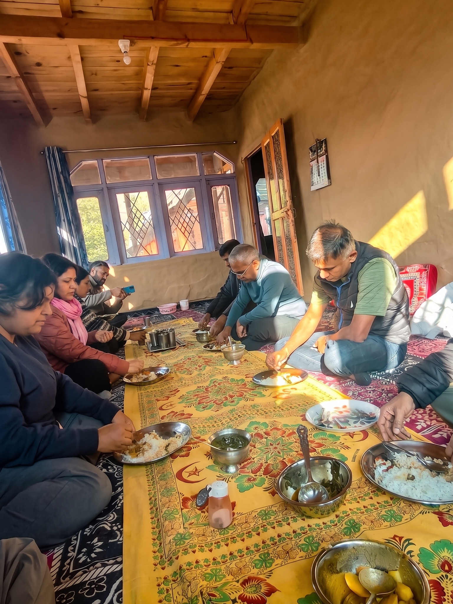 A group of people sitting cross-legged on a traditional patterned floor rug inside a wooden-ceilinged Kashmiri home, sharing a communal meal served on a yellow decorative cloth.