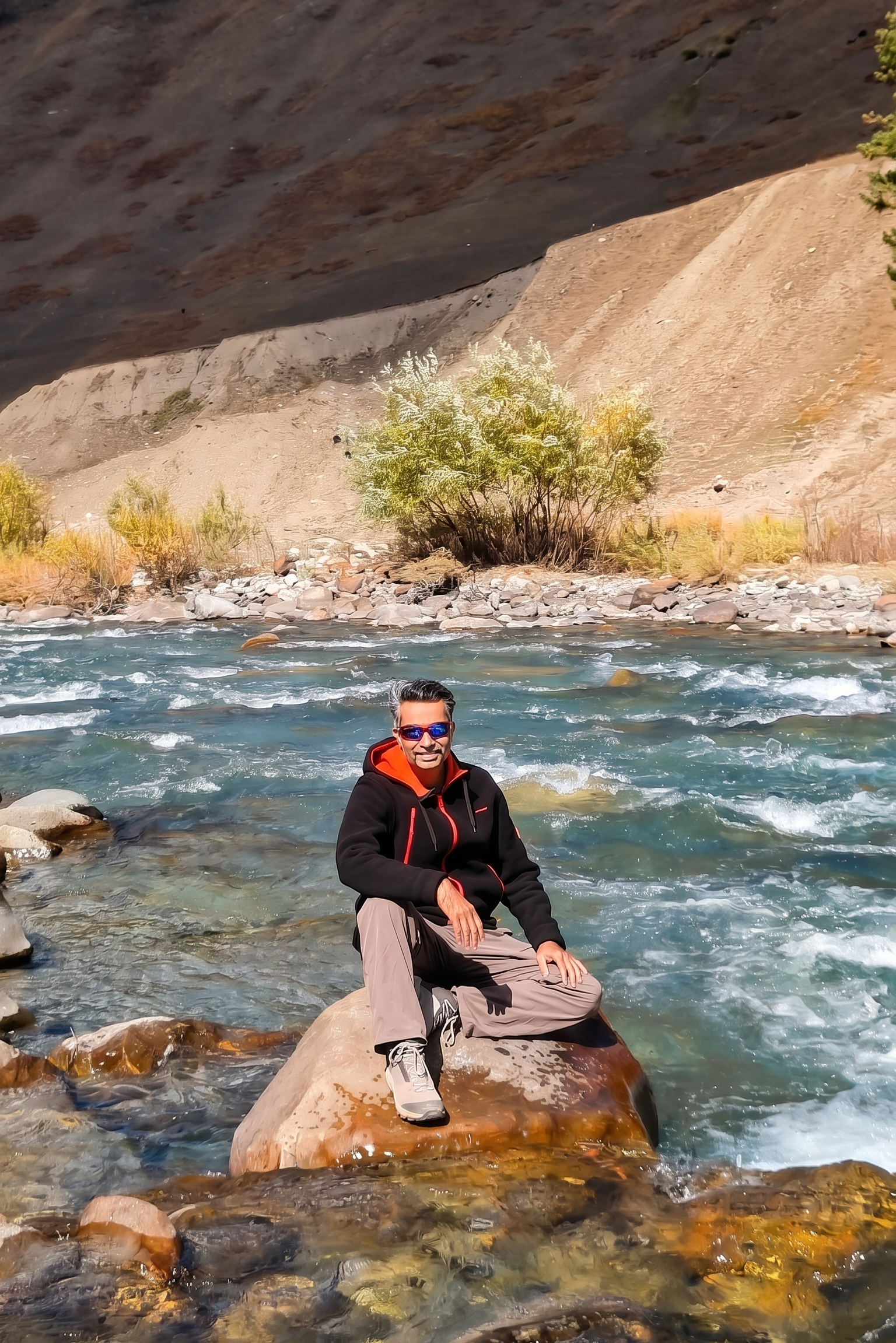 A man in a black jacket and sunglasses sitting cross-legged on a large boulder in the middle of a rushing turquoise mountain river in Gurez, with a steep valley hill in the background.