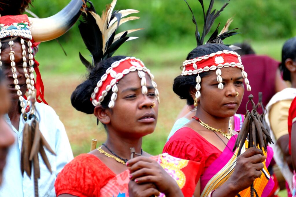 Tribal women in traditional Bastar attire wearing shell and feather headgear during a cultural performance.