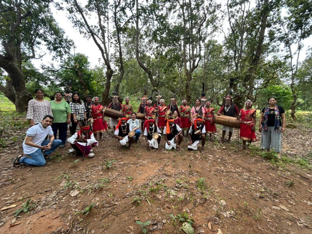 A group of tourists posing for a photo with indigenous Bastar tribal dancers wearing traditional red attire and horned headgears in a forested area.