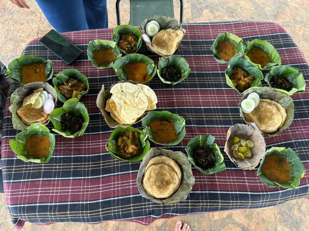 An overhead view of a traditional Bastar meal served in eco-friendly leaf bowls (dona) on a checkered tablecloth, featuring local curries, dals, flatbreads, puris, and salads.