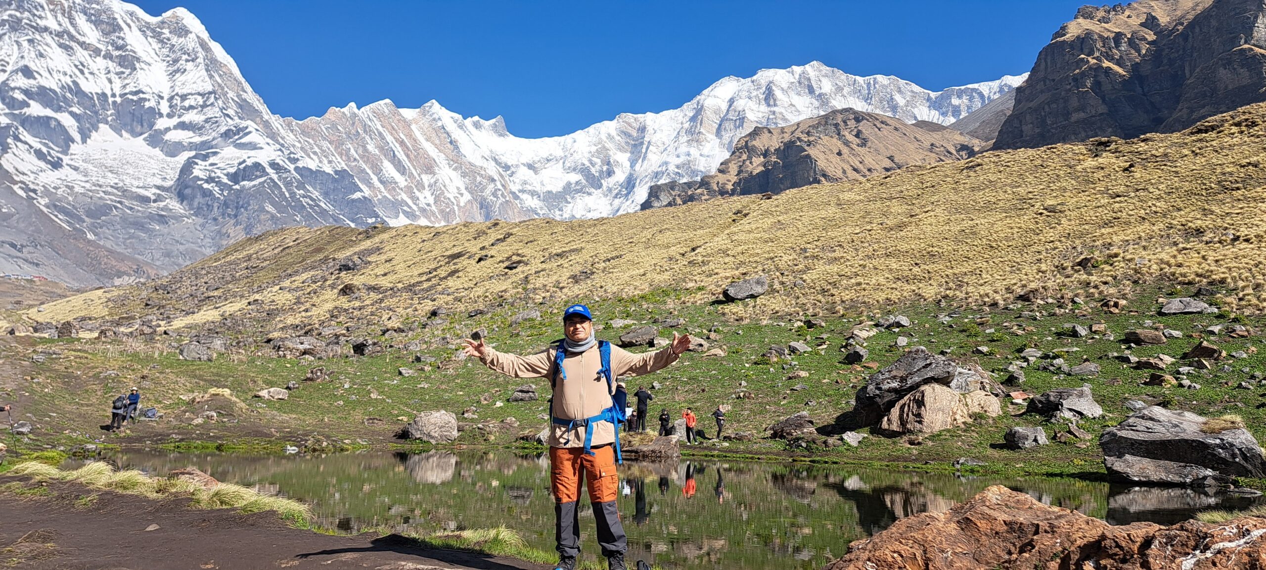 Trekker standing beside the alpine pond at Annapurna Base Camp with the Annapurna Amphitheatre and snow-covered Himalayan peaks in Nepal.